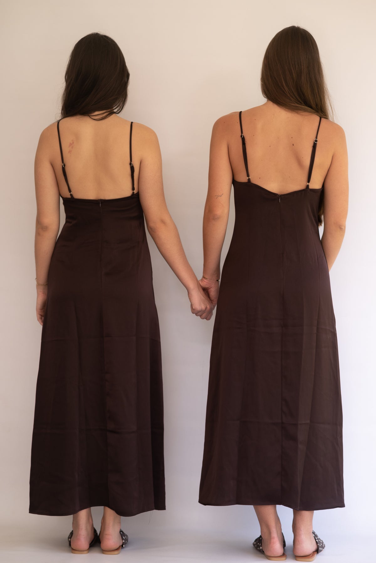 Two women wearing chocolate brown satin slip dresses holding hands against a plain background