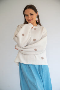 Woman wearing a white sweater with heart patterns and a blue skirt against a plain background