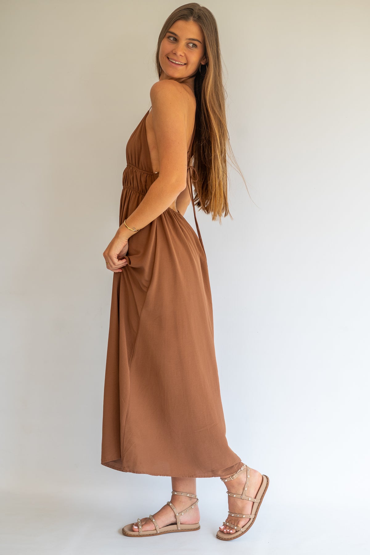 A woman modeling a sleeveless, brown maxi dress with a side cutout, paired with strappy flat sandals, photographed against a plain white studio background.