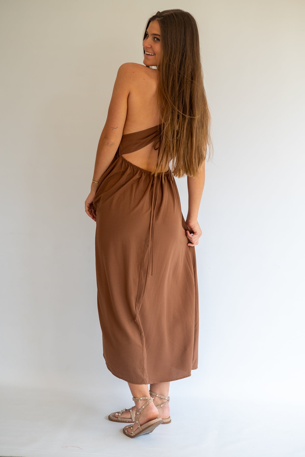 A woman modeling a brown backless wrap maxi dress, shown from the back against a plain white studio background.