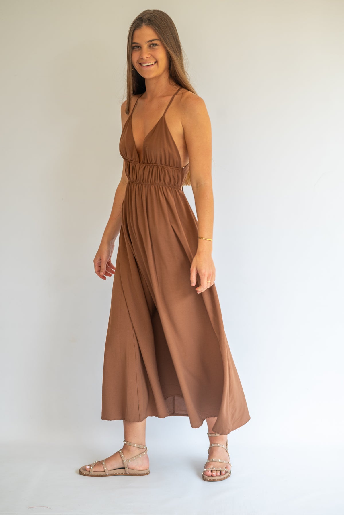 A woman modeling a sleeveless brown midi dress with a gathered waist and flowing skirt, paired with strappy flat sandals, against a plain white studio background.