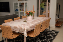 Load image into Gallery viewer, A dining room scene with a rectangular table covered in a beige and white damask-patterned tablecloth, surrounded by woven tan chairs. A pink vase with orange flowers and two tall taper candles sit on the table.
